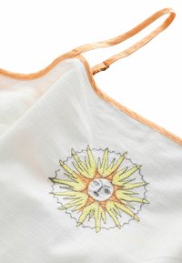 Biela látka s broskyňovými saténovými lemami predstavuje slnečný motív vyšívaný v žltej a oranžovej farbe s tvárou, obklopený špicatými lúčmi.