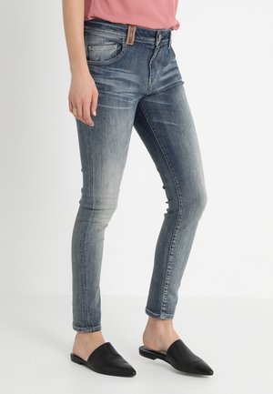 Jean skinny en denim bleu délavé avec un léger effet usé, cinq poches et une étiquette en cuir brun à la taille au dos. Mules noires portées.