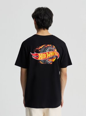 Persona con capelli neri corti che indossa una maglietta nera con un logo di pneumatico Hot Wheels infuocato sulla schiena, in piedi su uno sfondo semplice.