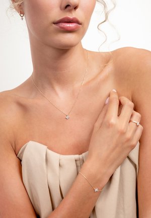 Goudkleurige ketting met een stervormige hanger, bijpassende armband en ring, allemaal met kleine heldere edelsteenaccenten. Glad huidkleurig stoffen achtergrond.