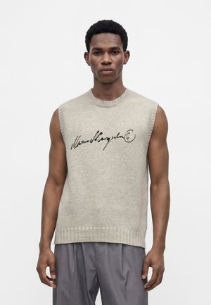 Homme portant un pull en maille beige sans manches avec un texte en écriture noire et un pantalon gris ample, debout sur un fond blanc uni.