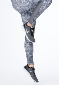 Šedé legíny se vzorem a geometrickými designy v kombinaci s černými sportovními botami, které mají bílou podrážku a výrazné logo swoosh.