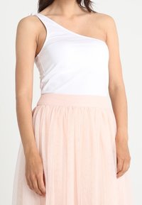 Witte one-shoulder top van rekbaar materiaal, gecombineerd met een zachtroze tule rok met een gerimpelde taille en gelaagde structuur.