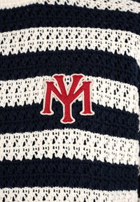 Logotipo bordado en rojo de NY sobre tela de punto texturizada con rayas horizontales en azul marino y blanco.