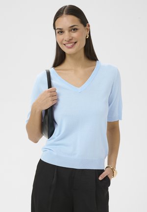 Jonge vrouw lacht, draagt een lichtblauw V-hals shirt met korte mouwen en zwarte broek, houdt een zwarte schoudertas vast en draagt gouden accessoires.