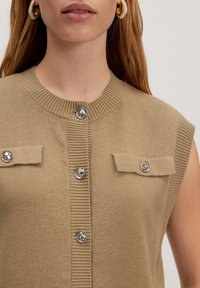 Beige ärmelloses Oberteil mit ribbeligem Halsausschnitt, zwei Klappentaschen und silbernen Knopfakzenten auf der Vorderseite. Glatte Stricktextur.