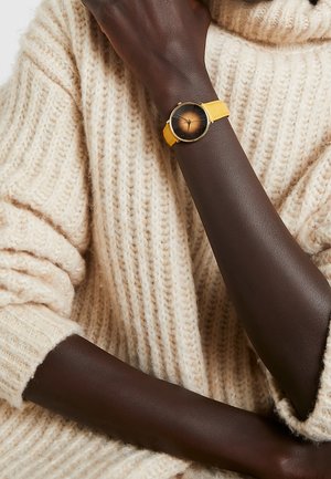 Personne à la peau foncée portant un pull en maille crème avec une montre à bracelet jaune affichant un cadran rond dégradé de marron à son poignet.