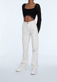 Person iført sort kort mesh langærmet bluse, hvide højtaljede cargo-bukser og hvide sneakers, stående på ensfarvet baggrund.