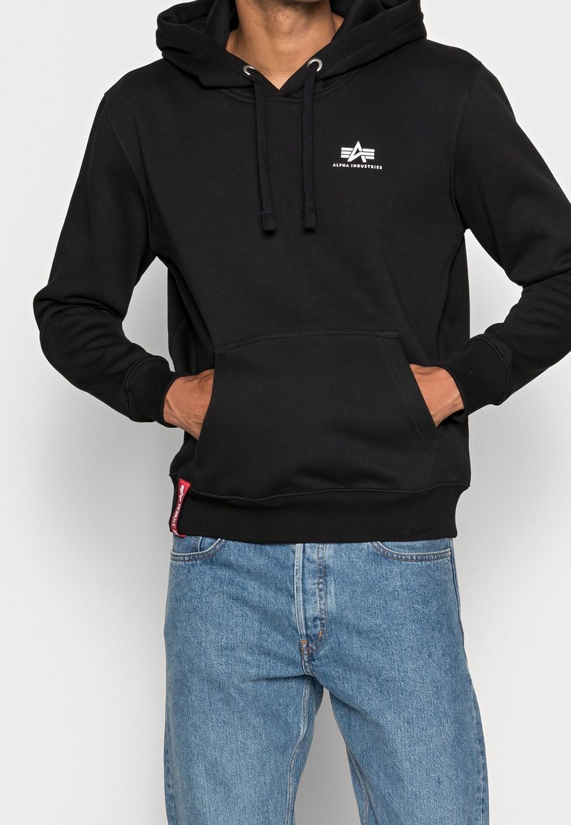 Persona con sudadera negra de Alpha Industries con bolsillo frontal y jeans azules, manos metidas dentro del bolsillo.