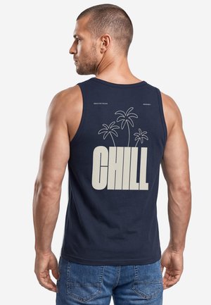 Man wearing a navy blue tank top with palm tree outlines and large text "CHILL" on the back, paired with blue jeans.