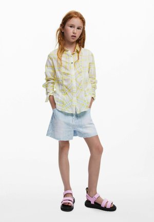 Cămașă cu nasturi albă cu carouri galbene, combinată cu o fustă din denim deschis. Încălțămintea include sandale roz cu tălpi și bretele negre.