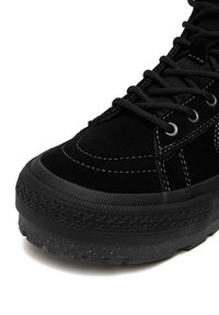 Melna zamšādas sporta apavu ar biezas faktūras gumijas zoli, melnām šņorēm un baltām šuvju detaļām uz gluda un raupja materiāla.