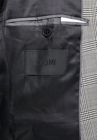 Svart tyginteriör med en dold ficka och knappstängning. Lapp som läser "KIOMI" sydd på tyget, intill ett texturerat rutmönster.