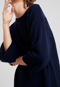 Granatowy sweter w oversize'owym stylu z szerokimi, rękawami do łokci i miękką teksturą. Posiada dekolt łódkowy oraz subtelne detale szwów.