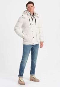 Giacca a vento bianca con cappuccio, dotata di cerniere e bottoni neri, abbinata a jeans blu e stivali color cammello con lacci e suole in gomma.