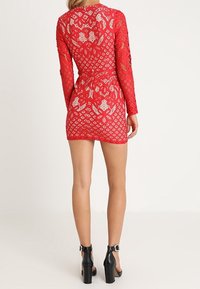 Robe en dentelle rouge à manches longues, silhouette ajustée, avec motifs floraux et dos nu. Portée avec des talons noirs à bride à la cheville.