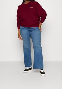 Bordeauxrode sweatshirt met ronde hals, gecombineerd met lichtblauwe flared jeans. De outfit wordt compleet gemaakt met zwarte sneakers met witte veters.