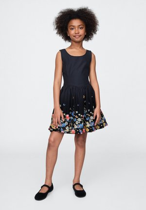 Marineblauwe jurk met een aangesloten lijfje en een uitlopende rok. De zoom is versierd met bloemenprints in verschillende kleuren. Gecombineerd met zwarte platte schoenen.