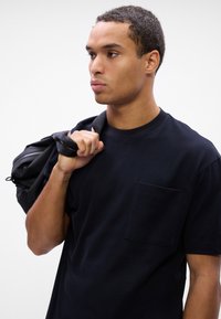 Μαύρη κοντομάνικη μπλούζα με στρογγυλή λαιμόκοψη και τσέπη στο στήθος. Το υφάσμα φαίνεται λείο και μαλακό. Το υποκείμενο κρατά μια μαύρη τσάντα στον έναν ώμο.