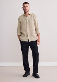 Camicia a righe color tannino con bottoni e maniche arrotolate, abbinata a pantaloni neri e mocassini. Design semplice, tessuto strutturato e colori neutri.