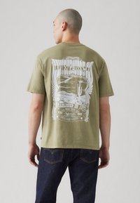 Olive green short-sleeve t-shirt featuring a large white graphic design on the back, with vintage lettering and desert imagery.