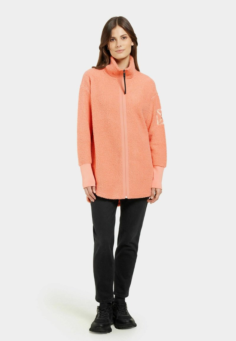 Korallfarbene, flauschige Zip-Jacke mit hohem Kragen, langen Ärmeln, kontrastierenden Bündchen und einem Frontreißverschluss. Getragen mit schwarzen Hosen und schwarzen Schuhen.