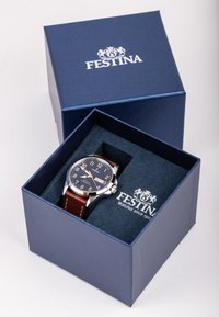 Festina laikrodis mėlyname dėkle, su ruda odine juosta, sidabriniu korpusu, tamsiai mėlynu ciferblatu, baltais skaičiais ir prekės ženklu viduje.