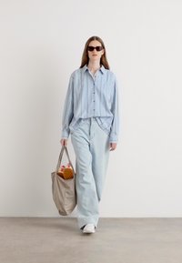 Light blue striped button-up shirt, loose light blue jeans, white sneakers, and a beige tote bag containing shoes. Model wears black sunglasses.