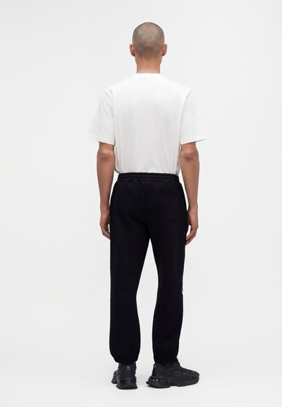 Homme à la tête rasée portant un t-shirt blanc uni, un pantalon de jogging noir et des baskets noires, debout de dos sur un fond blanc.