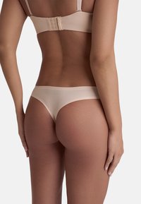 Perizoma beige chiaro realizzato in un materiale morbido e elastico, caratterizzato da copertura minima, design senza cuciture e senza hardware visibile.