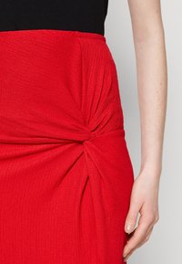 Jupe rouge texturée avec un détail en design torsadé à la taille et un haut lisse. Le matériau semble extensible.