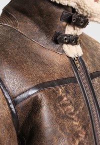 Veste en cuir marron avec col en peau lainée, dotée d'une fermeture éclair et de deux sangles réglables. Texture lisse avec des accents en cuir foncé contrastants.