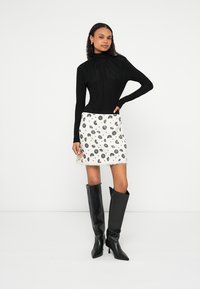 Μαύρο τοπ με ψηλό λαιμό και μακριά μανίκια, λευκή μίνι φούστα με μαύρο λουλουδάτο μοτίβο, υφασμάτινη υφή, μαύρες μπότες με τακούνι μέχρι το γόνατο, σε όρθια στάση.