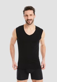 Black sleeveless V-neck shirt in a fitted design, made of smooth fabric, showcased on a male model with short hair and beard.