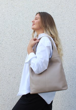 Borsa a tracolla in pelle beige con una texture morbida, presenta un'ampia apertura e una spallina robusta, portata da una persona in camicia bianca.