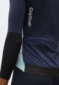 Κοντινό πλάνο ενός ατόμου που φορά μπλε σκούρο και μαύρη φανέλα ποδηλασίας με το λογότυπο "GripGrab" στο πλάι και στην πίσω τσέπη.