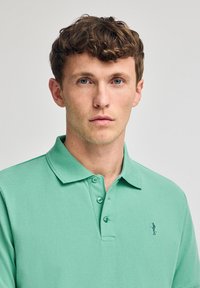 Camisa polo verde menta con cuello, tres botones y un pequeño logo en la parte izquierda del pecho. Textura suave, diseño casual y estilo ajustado.