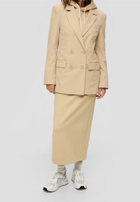 Žena nosící béžové pruhované dvouřadé sako, béžovou mikinu s kapucí, béžovou midi sukni, bílé ponožky a bílé tenisky.