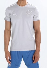 Šedé sportovní tričko s krátkým rukávem a bílým logem Adidas, emblémem týmu a třemi pruhy na ramenou. Nosené s modrými sportovními šortkami.