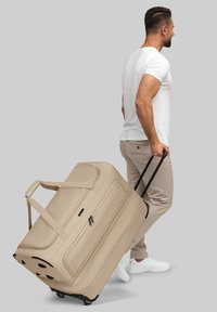 Uomo in t-shirt bianca e pantaloni beige che trascina una grande borsa a rotelle beige contro uno sfondo grigio uniforme.
