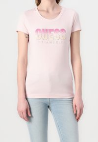 Kobieta w jasnoróżowej koszulce z krótkim rękawem z logo "GUESS Los Angeles" w gradientzie różowo-żółtym oraz jasnoniebieskich dżinsach, z widocznym tatuażem na ramieniu.