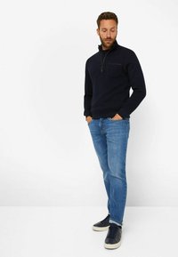 Marineblauer Pullover mit halbem Reißverschluss und Fronttasche, kombiniert mit hellblauen Jeans und schwarzen Sneakers. Glatte Stoffoberfläche.