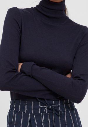 Person trägt einen enganliegenden marineblauen Rollkragenpullover und marineblaue Hose mit weißen vertikalen Streifen und gebundenem Gürtel, Arme verschränkt.