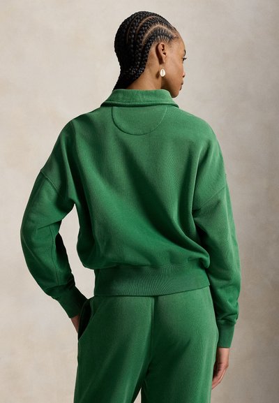Femme aux cheveux tressés portant un haut à manches longues vert et un pantalon vert assorti, debout de dos avec la tête tournée sur le côté.