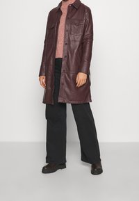 Manteau en cuir bordeaux avec fermeture à boutons, deux poches poitrine et col à revers, associé à un pull rose et un pantalon noir à jambes larges.