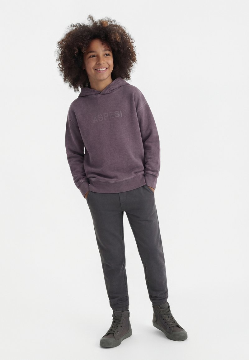 Bambino con capelli ricci sorridente, indossa una felpa con cappuccio viola con la scritta "ASPESI", pantaloni scuri e sneakers alte grigie, in piedi con le mani nelle tasche.