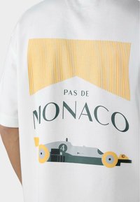 Hvid bomuld t-shirt med gul stribet grafik, der har teksten "PAS DE MONACO" og en stiliseret racerbilsillustration i grå og gul.