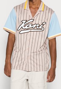 Uomo che indossa una camicia a maniche corte beige a righe con maniche azzurro chiaro e il logo "Kami" sul petto, abbinata a pantaloni bianchi.