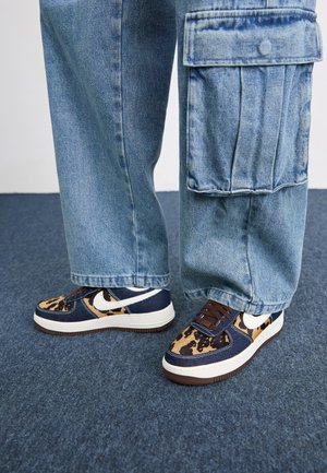 Personne portant un pantalon cargo large en denim bleu et des baskets à motifs avec le swoosh blanc de Nike, debout sur un tapis bleu.