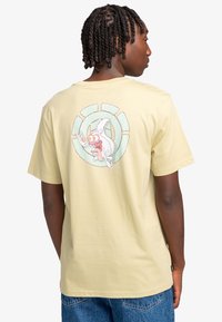 Lichtgele katoenen t-shirt met een grafisch ontwerp van een karakter met grote ogen en een cirkelpatroon op de achterkant. Korte mouwen.